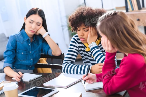 Preview: bored multiracial students sitting at table while doing homework together
