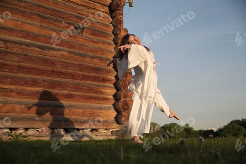 Preview: A woman in white attire dances gracefully next to a wooden building at sunset. This image reflects