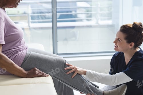Preview: Diverse senior female patient exercising leg and female doctor advising in hospital room