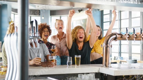 Preview: Group Of Young Male And Female Friends Celebrating Whilst Watching Game On Screen In Sports Bar