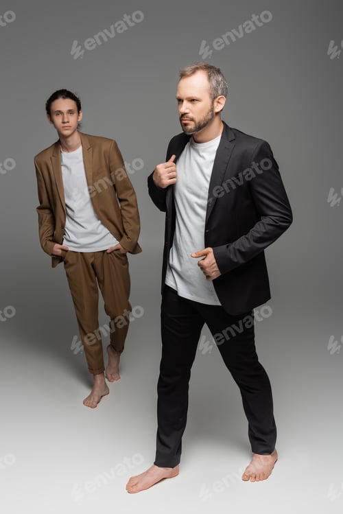 Preview: full length of well dressed man walking with bare feet next to stylish teenage boy on grey