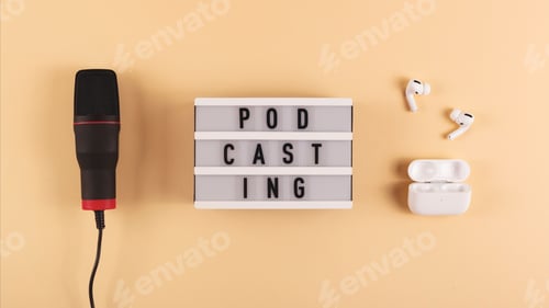 Preview: Podcasting lettering next to microphone and wireless headphones at workplace on beige background.