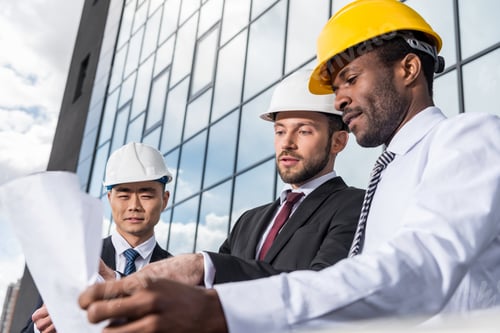 Preview: Multiethnic group of professional architects in helmets working with blueprint outside modern