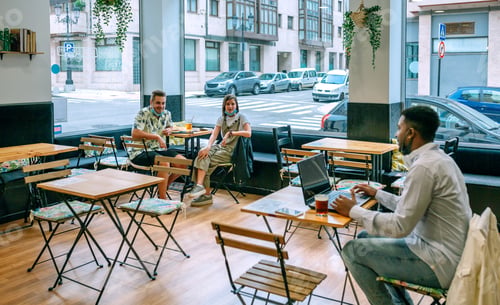 Preview: Young man teleworking in coffee shop talking to friends maintaining social distance