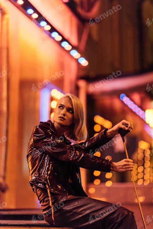 Preview: attractive young woman in leather jacket sitting on street at night under yellow light and holding