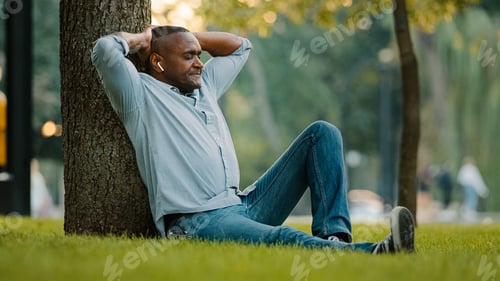 Preview: Calm relaxed resting middle-aged male listening music with earphones sitting on green grass in park