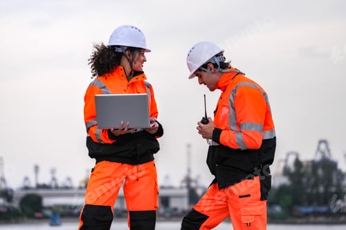 Preview: Two engineers wearing gear review plans on a laptop at a shipyard,