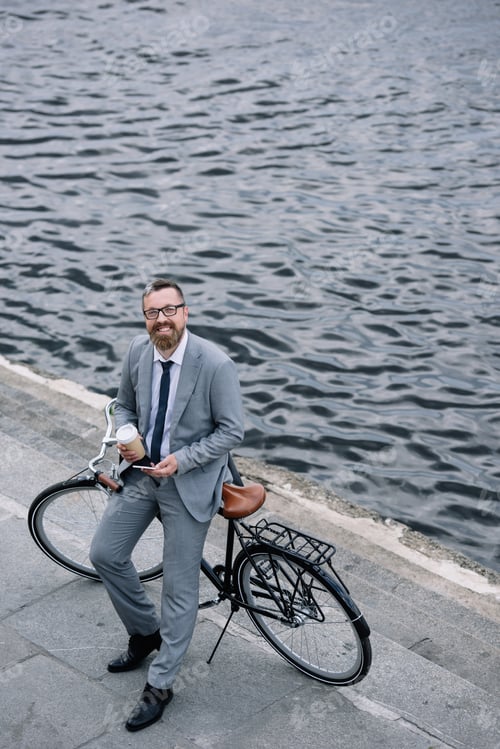 Preview: businessman with coffee to go and smartphone standing with bicycle on quay