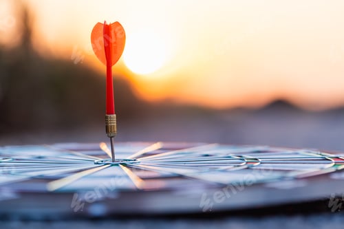 Preview: Close up red dart arrow hitting target center dartboard on sunset background.