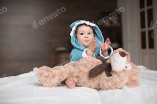 Preview: Happy baby boy in blue robe playing with teddy bear on bed