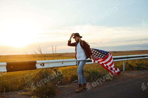 Preview: 4th of July. American Flag. Traveler with flag of America. Man in hat, backpack, a shirt and jeans.