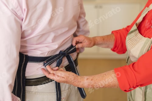 Preview: Mid section of senior caucasian couple tying apron in kitchen