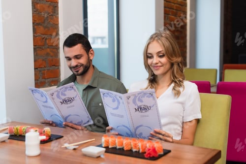 Preview: happy couple choosing meal from menu near blurred sushi rolls in restaurant