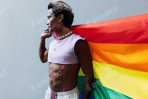 Preview: Young gay standing with a pride flag at grey wall and looking away