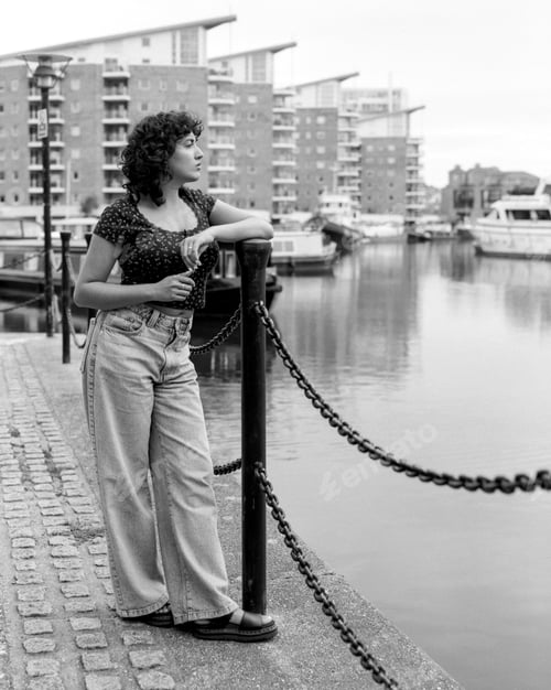 Preview: Young woman leaning on pole contemplating canal in monochrome analog medium format film