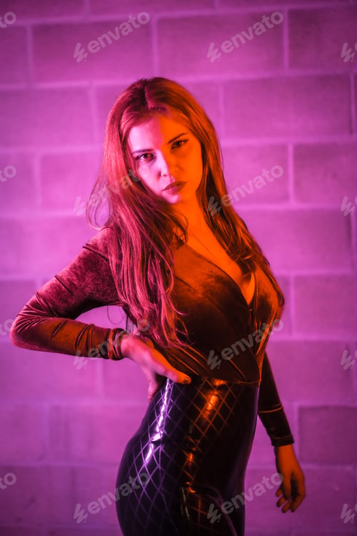 Preview: brown and black tight suit on a brick wall, posed at night illuminated with pink light