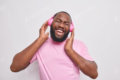 Preview: Horizontal shot of happy handsome man keeps hands on stereo headphones enjoys favorite music tilts h