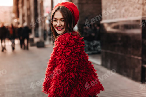 Preview: Fashionist girl in shaggy red coat and bright headdress smiles, walking along busy city street