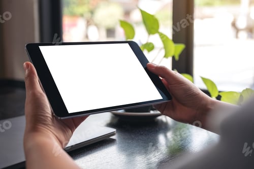 Preview: hands holding black tablet pc with blank white desktop screen with green leaves on wooden table