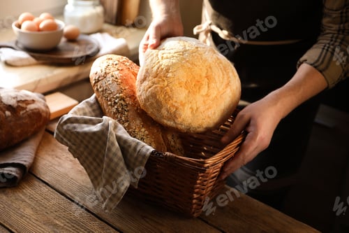 Preview: Man holding wicker basket with different types of bread at wooden table indoors, closeup