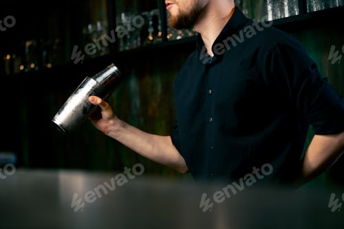 Preview: Professional bartender in black shirt holds in hands steel shaker Blurred bottles on shelves in the