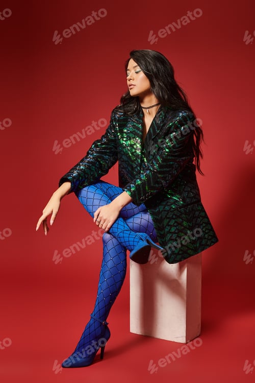 Preview: trendy asian woman in green jacket with sequins and blue pantyhose sitting on red backdrop