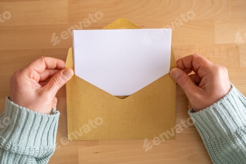 Preview: Woman’s hands opening brown envelope with blank white letter on wooden table, ready for design.