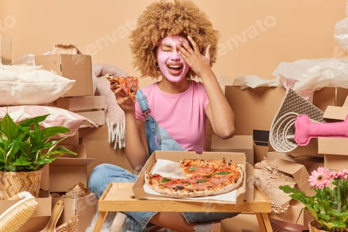 Preview: Indoor shot of positive woman wears beauty facial mask covers face with palm eats delicious pizza ha