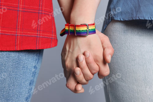 Preview: LGBT concept. Women in rainbow wristbands holding hands on light grey background, closeup
