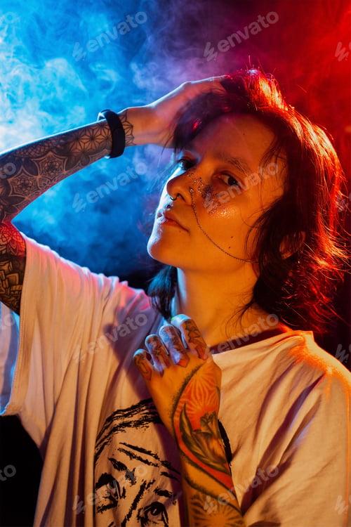 Preview: Stylish young tattooed woman with face piercing posing on dark background with smoke in neon lights.