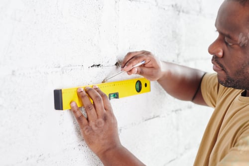 Preview: African man with pencil drawing line on white painted brick wall while using level measuring handtoo