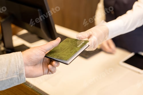 Preview: Gloved hand of young female receptionist passing passport to guest of hotel over reception counter