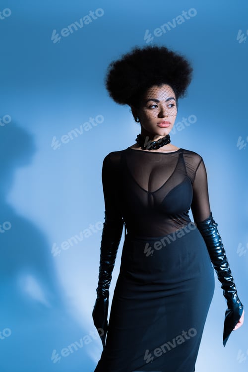 Preview: african american woman in sexy outfit and black veil looking away while posing on blue