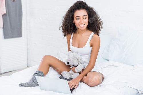 Preview: curly young woman holding teddy bear and using laptop in bed during morning time at home