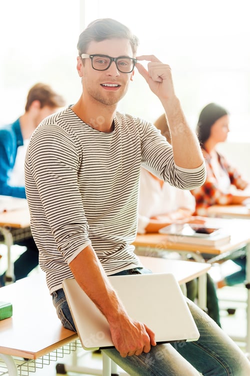 Preview: Confident student. Smiling young man holding laptop and adjusting eyewear while leaning at the desk