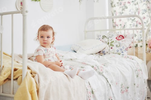 Preview: Baby girl in cute dress siting at bed playing with toys by the home. White vintage childroom