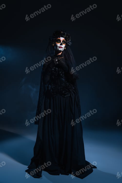 Preview: full length of woman in black witch dress and halloween makeup on dark background with blue light