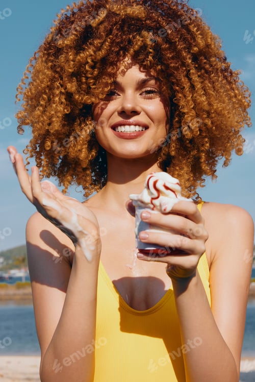 Preview: Young woman with curly hair enjoying ice cream at the beach Bright yellow swimsuit with a sunny blue