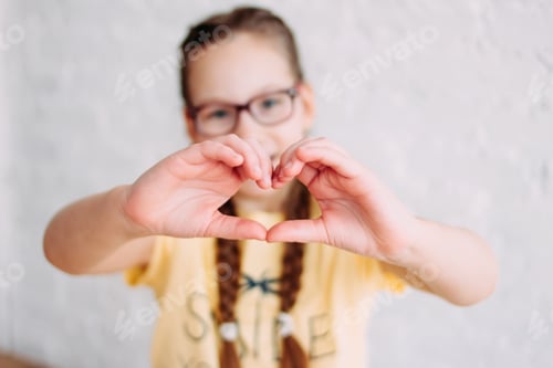 Preview: Happy tweens girl in yellow t-shirt making heart by hands on the white wall background