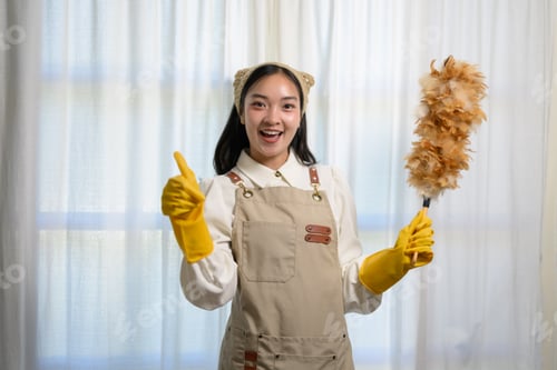 Preview: Young smiling maid wearing yellow gloves and an apron is holding a feather duster and showing thumbs