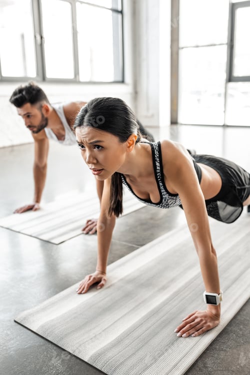 Preview: concentrated young woman and man doing push ups on yoga mats in gym