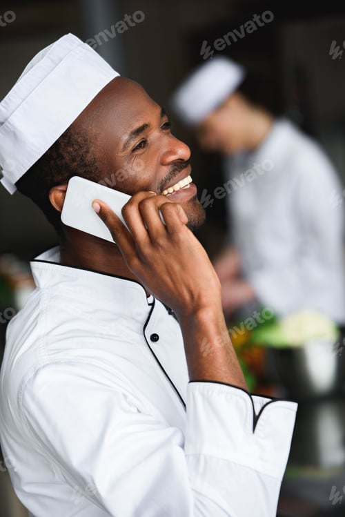 Preview: side view of smiling african american chef talking by smartphone at restaurant kitchen