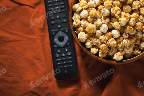Preview: Wooden bowl with sweet popcorn and TV remote on orange bedding. Top view with copy space
