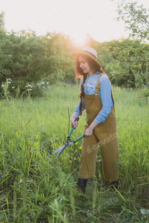 Preview: Cute young woman gardener in a straw hat cuts grass and weeds in an old garden with pruning shears