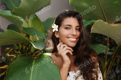 Preview: Tanned green-eyed lady in white top and with small flower in her long hair with charming smile look