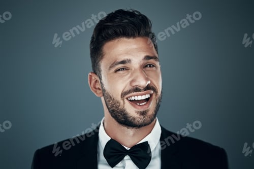 Preview: Charming young man in bow tie looking at camera and smiling while standing against grey background