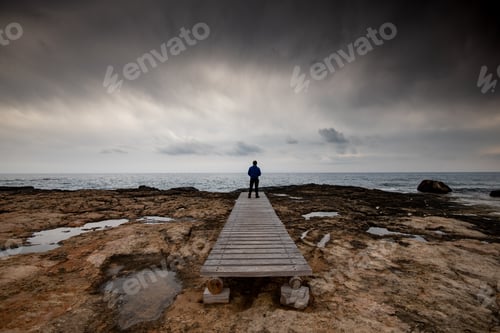Preview: Man standing alone at the edge of rocky beach in Stormy Weather