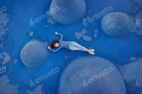 Preview: woman in blue jeans and a shirt is lying on a blue background on a sports field or in a park. a