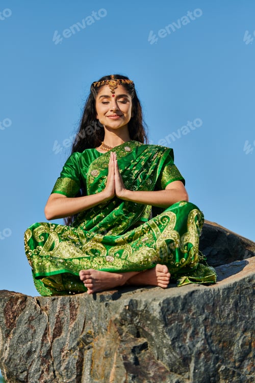 Preview: smiling indian woman in sari meditating while sitting on stone with blue sky on background