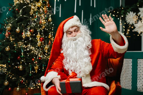 Preview: Santa Claus holding a gift and waving looks at the camera.